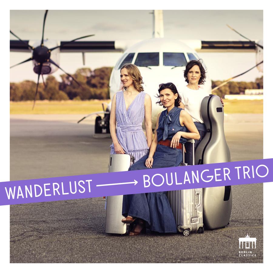 Release-Cover „Wanderlust“ des Boulanger Trio. Drei Musikerinnen mit Instrumentenkoffern vor einem Flugzeug, überlagert von violettem Titelband; warme Tageslichtfarben, frontale Gruppenaufnahme und reisefreudige, helle Atmosphäre.