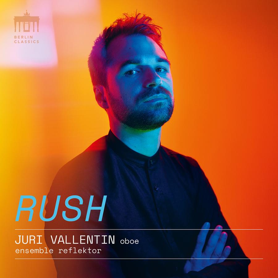 Release-Cover „Rush“ von Oboist Juri Vallentin mit ensemble reflektor. Farbiges Porträt mit verschränkten Armen vor orangefarbenem Hintergrund und blauem Licht auf dem Gesicht; kontrastreiche, moderne Studioaufnahme mit sichtbarer Typografie.