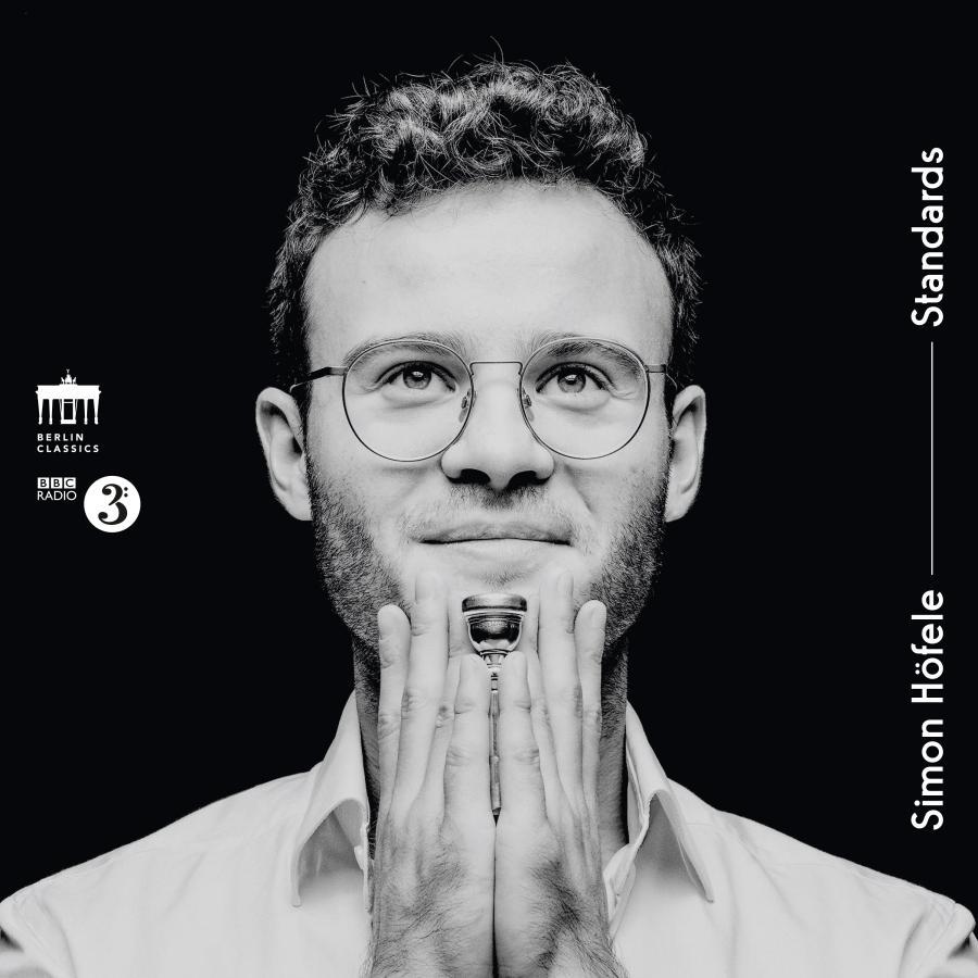 Release-Cover „Standards“ von Trompeter Simon Höfele. Schwarz-Weiß-Porträt mit Brille, hält Trompetenmundstück ans Kinn; schwarzer Hintergrund, frontale Nahaufnahme und klare, reduzierte Jazz-Ästhetik.