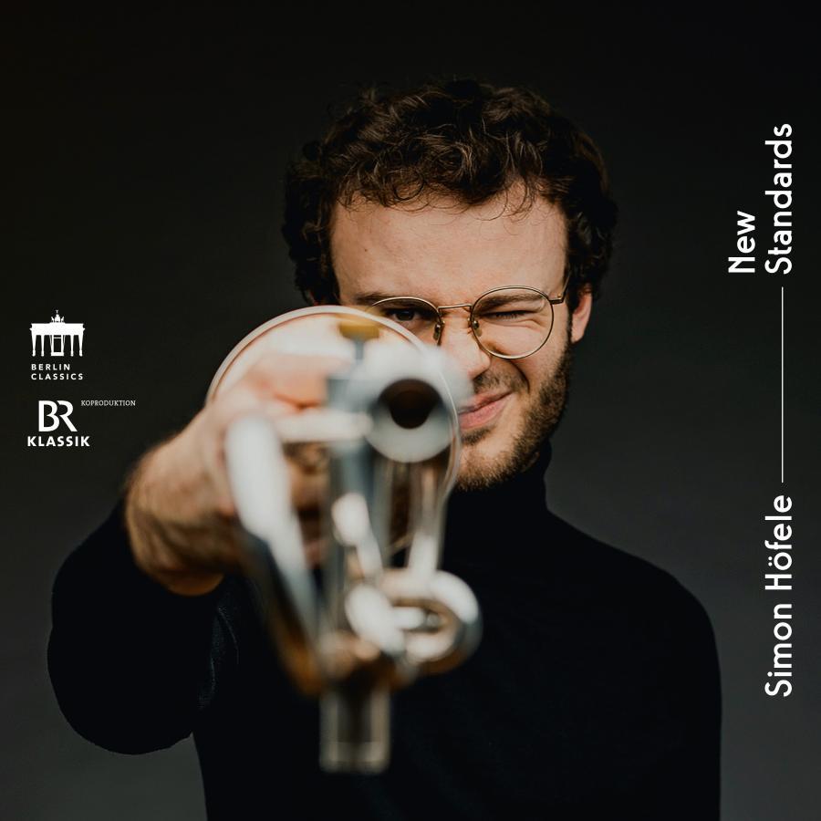 Release-Cover „New Standards“ von Trompeter Simon Höfele. Dunkles Porträt, wo Simon Höfele seine Trompete frontal in die Kamera hält; geringe Schärfentiefe, schwarzer Hintergrund und direkte, moderne Musikerinszenierung.