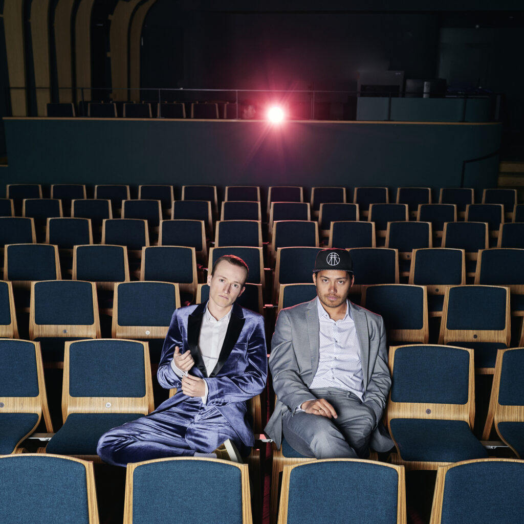Auf dem Bild sieht man das Dou Konstantin Reinfeld und Benyamin Nuss. Sie sitzen in einem Kino, tragen schöne Anzüge und wirken cool.