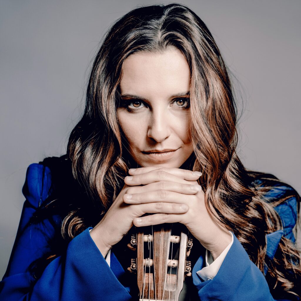 Porträt der Gitarristin Carlotta Dalia mit offenen Haaren. Sie trägt ein blaues Jacket und hat die Hände verschränkt auf dem Kopf einer Gitarre abgelegt.