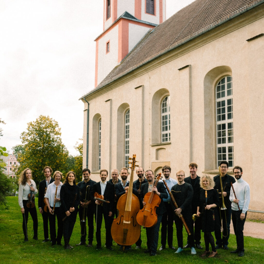 Das Ensemble il Gusto Barocco steht vor einer Kirche mit Instrumenten wie Cello, Kontrobass und Geige in den Händen.