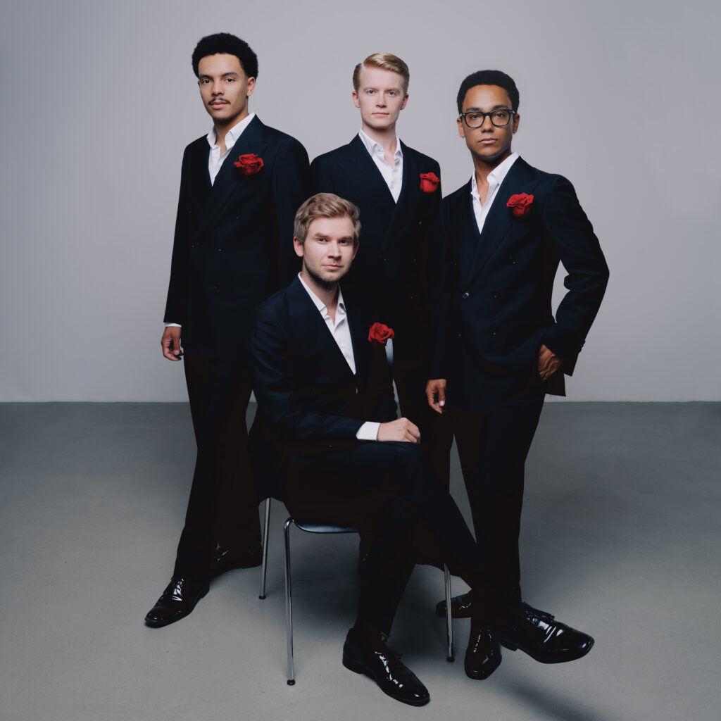 Zu sehen ist das A Capella Ensemble German Gents in schwarzen Anzügen mit weißem Hemd und rotem Einstecktuch. Sie sind sehr festlich gekleidet und schauen seriös in die Kamera. Der Bass Marcel Raschke sitzt auf einem Stuhl, während die anderen drei Mitglieder Thoma Jaron-Wutz (Tenor), Armin Horn (Tenor) und Raphael Riebesell (Bariton) hinter ihm stehen.
