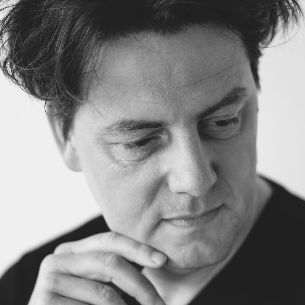 Portrait von Komponist Helge Burggrabe. Schwarz-Weiß-Foto in enger Nahaufnahme, er blickt nach unten und stützt das Kinn auf die Hand; weiche Grautöne, ruhige Lichtstimmung und kontemplative Wirkung.